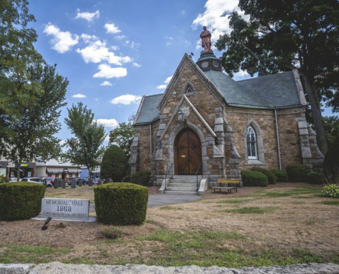 Granite Gothic Revival hall with arched wood double doors below a slate roof and turret statue.