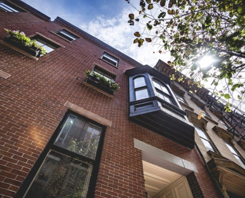 Brick rowhouse seen from below with a projecting clad oriel bay, arched sash, and window boxes.